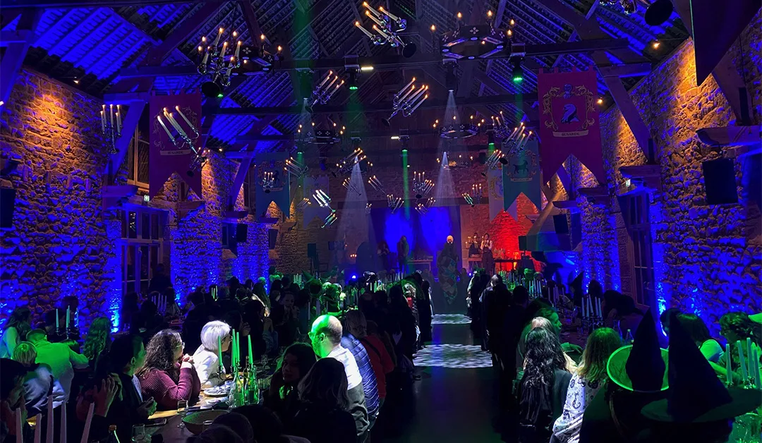 Imagen del salón de banquetes de la nueva escuela de magia - Le Rocher Portail. La decoración es absolutamente increíble, con candelabros suspendidos del techo, los escudos de armas de las diferentes familias, el juego de luces azules, verdes y rojas. La sala está llena.